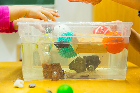 In einem großen, durchsichtigen Wassergefäß lassen Kinder verschiedene Gegenstände schwimmen und sinken.