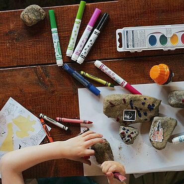 Kinderhände über einem Basteltisch mit Steinen, Stickern, Wassermalfarben und bunten Stiften.